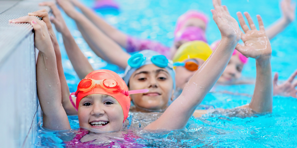 Children in their swimming lessons waving at the camera, they wear bright hats and goggles