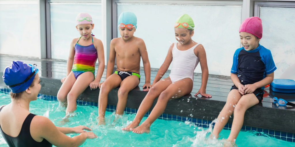 Children sit on the side of a swimming pool and listen to their swimming teacher