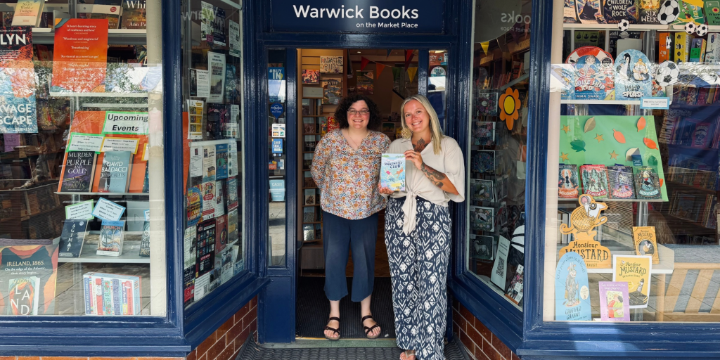 Two women stand outside a book shop and one of them is holding a book called The Underwater Club